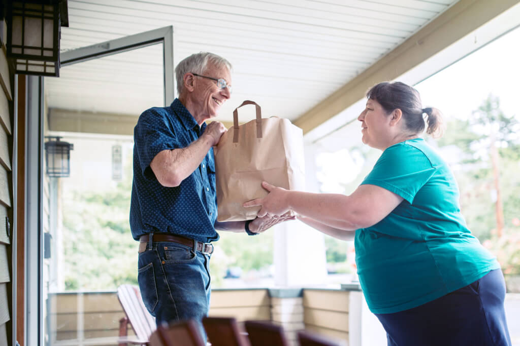 A kind and caring neighbor or friend delivers fresh produce from the grocery store to an elderly man at his home.  He receives the gift with a smile on his face, grateful for the help and assistance.  Horizontal image.