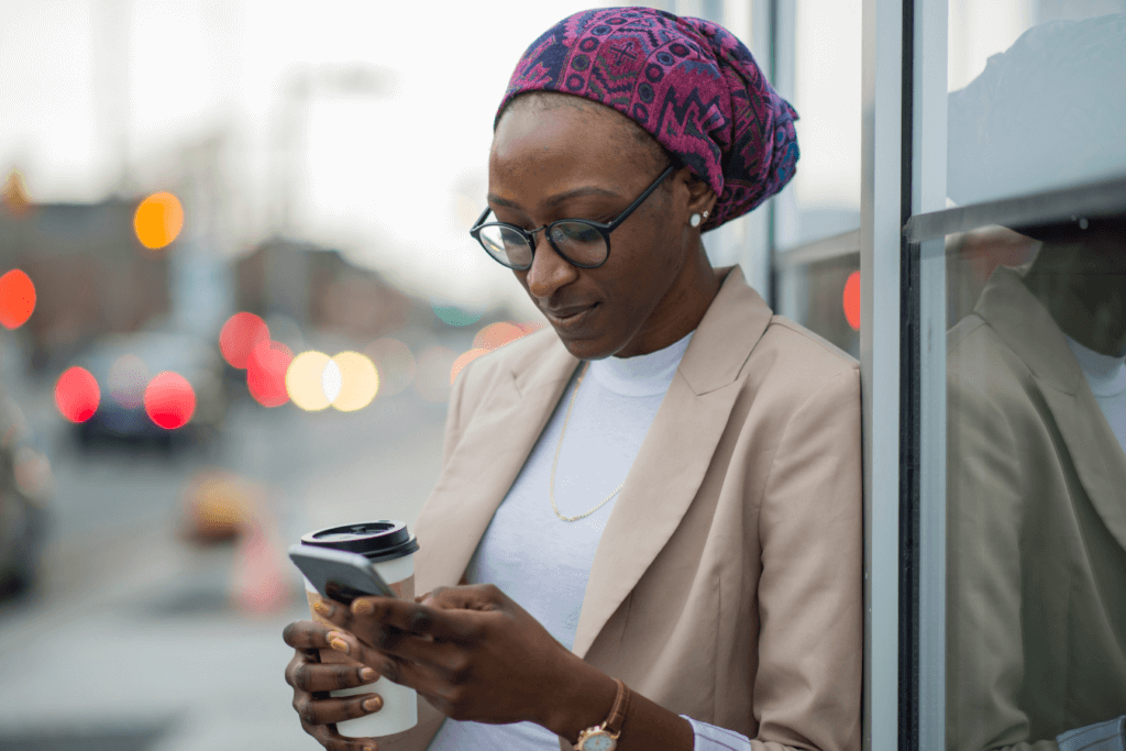 Black woman wearing glasses and a head wrap looking down at her phone and holding a cup of coffee outside a building