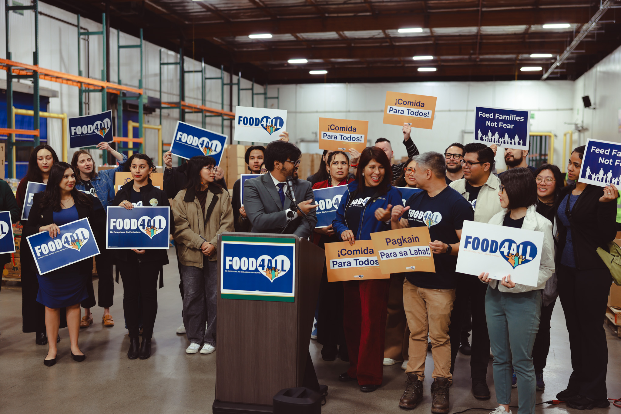Advocates stand together behind a podium in a food bank warehouse with signs that say "Food for all" in English, Spanish, and Tagalog.