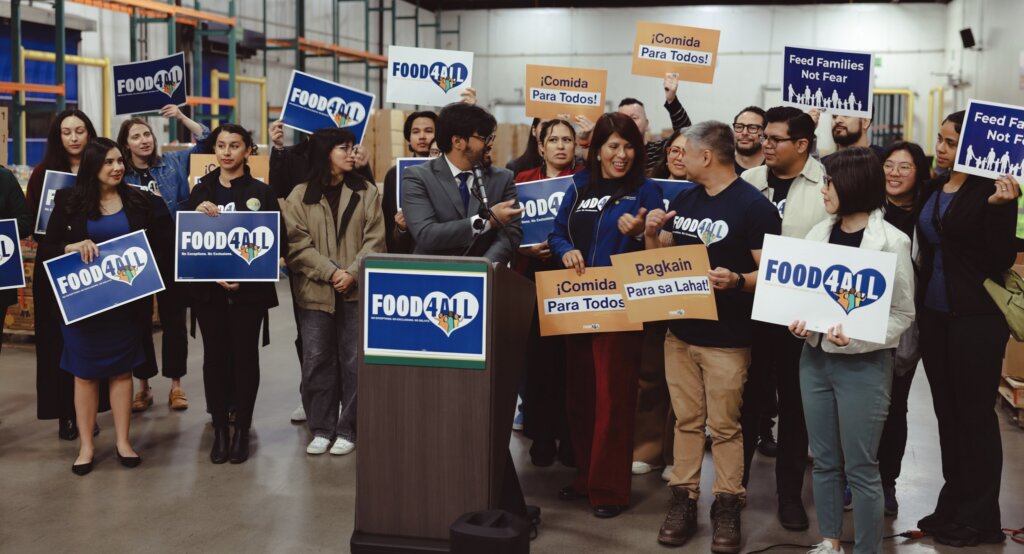 Advocates stand together behind a podium in a food bank warehouse with signs that say "Food for all" in English, Spanish, and Tagalog.