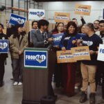 Advocates stand together behind a podium in a food bank warehouse with signs that say "Food for all" in English, Spanish, and Tagalog.